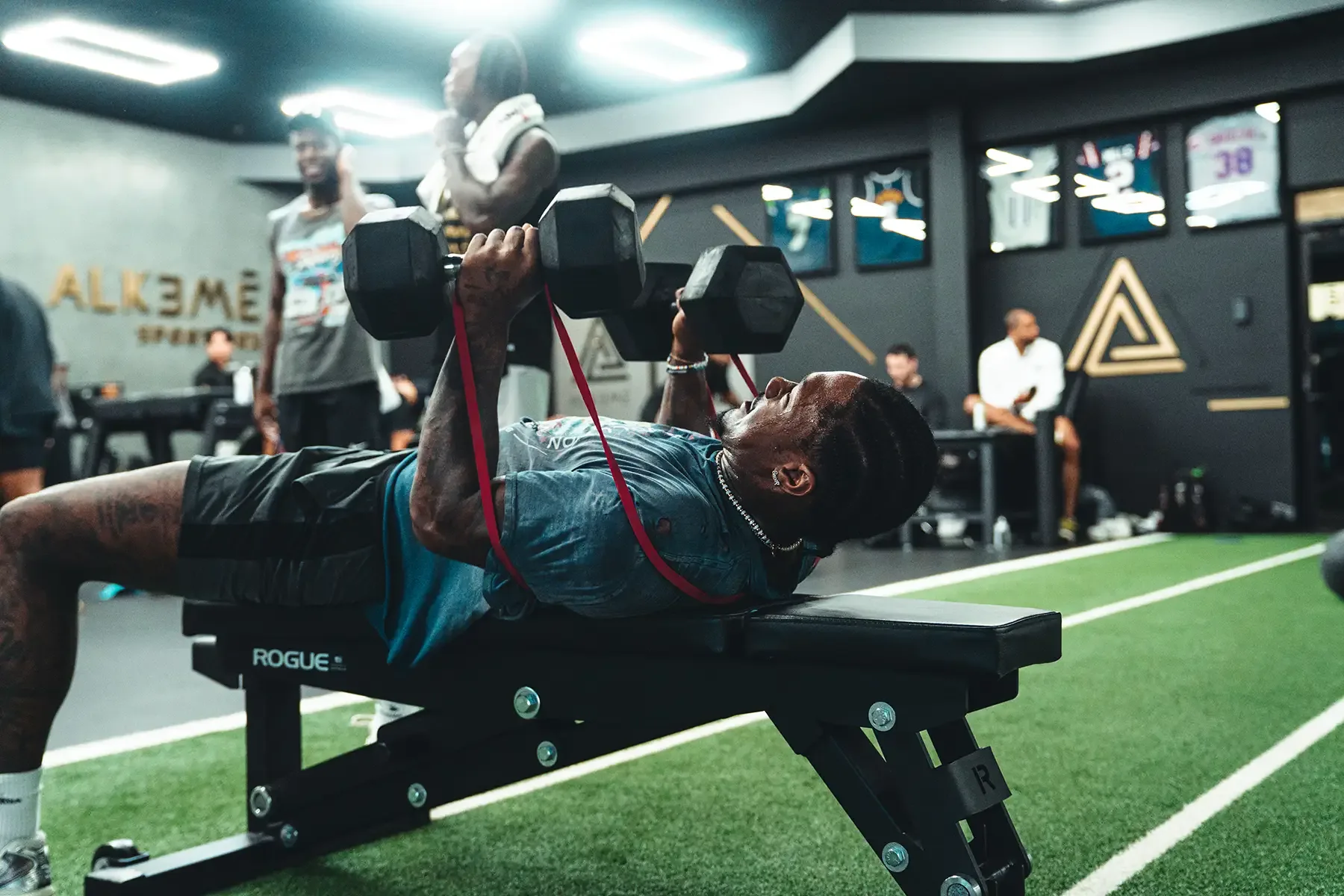 An athlete pressing dumbbells against band resistance on the turf at Alkemē Sports Rx in Davie, FL