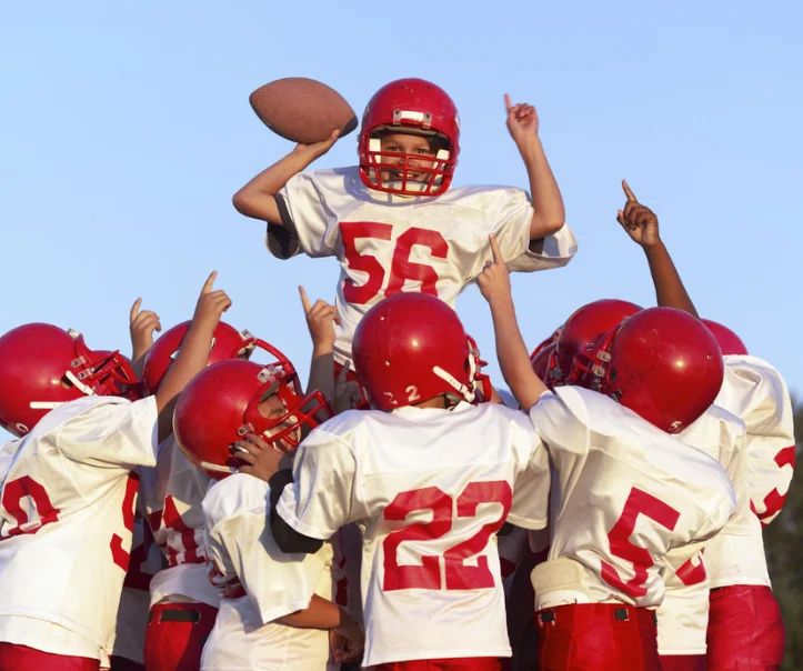 Youth football team celebrating with a player in a red jersey holding a football, surrounded by teammates in matching uniforms, emphasizing teamwork and sportsmanship.