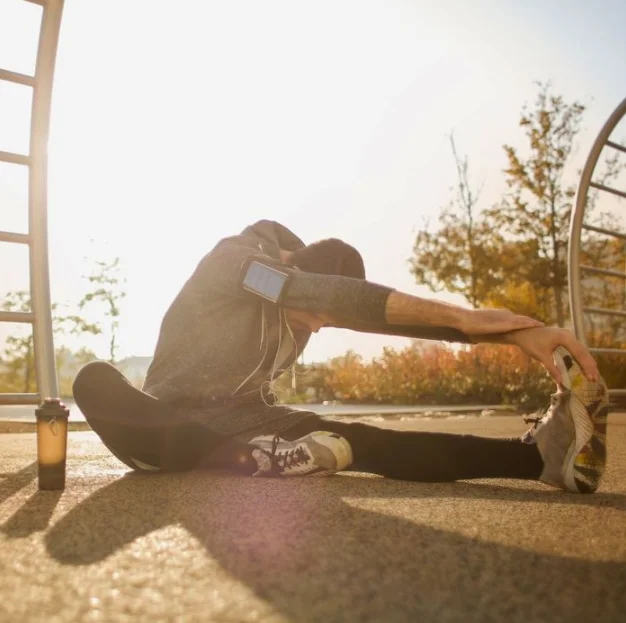Athlete stretching in outdoor training environment, emphasizing flexibility and core strength, with a water bottle nearby, reflecting sports performance training focus at Alkeme Sports RX.
